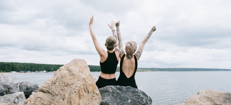 Two women sitting near the water with their hands in the air.